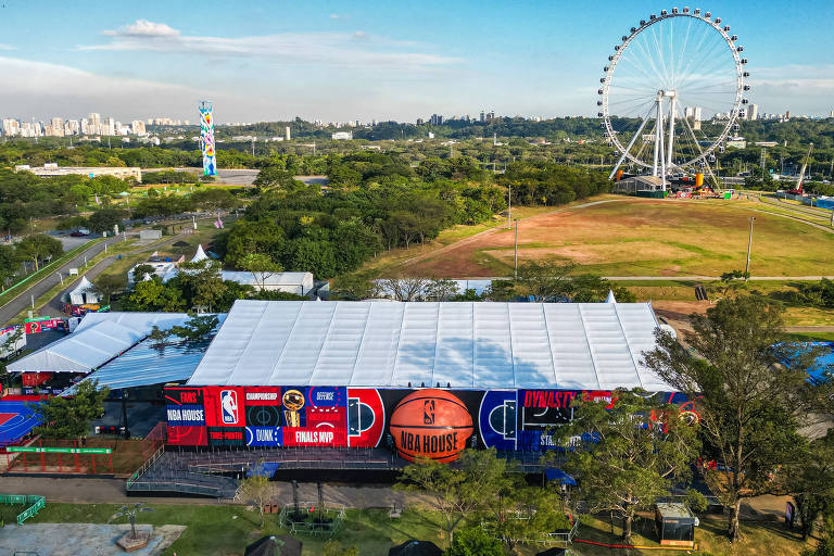A imagem mostra uma vista aérea de um evento da NBA em São Paulo, com uma grande estrutura coberta, decorada com logotipos da NBA e do evento. Ao fundo, há uma roda-gigante e uma área verde. O céu está claro e a cidade é visível ao longe, com prédios e árvores ao redor.