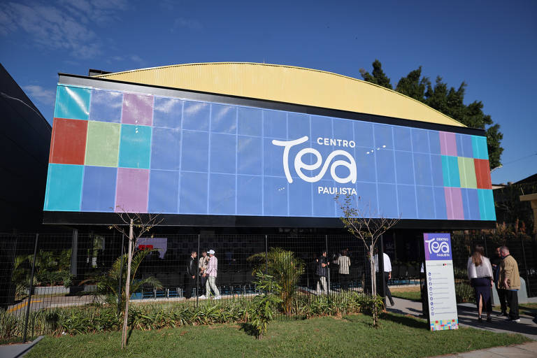 A foto em um ambiente externo mostra um jardim, com grama e árvores verdes, e uma placa azul com detalhes vermelhos, amarelos, azuis e rosas escrito "Centro TEA Paulista" em letras brancas.