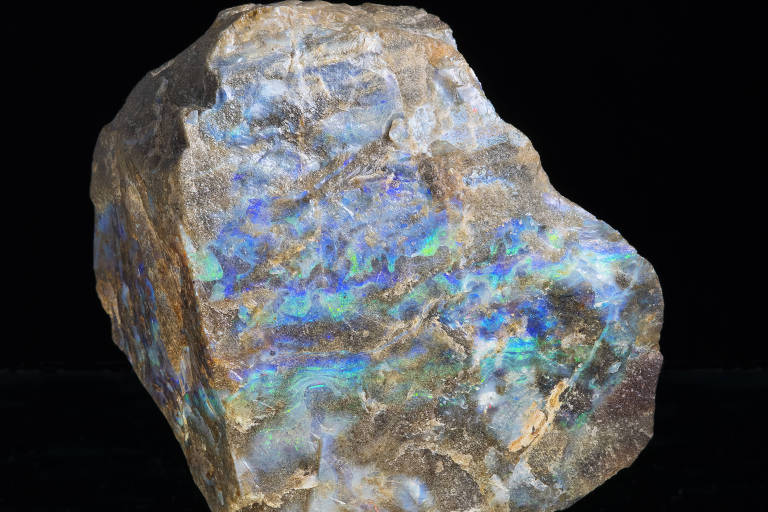 Uma pedra de labradorita, com uma superfície irregular e cores iridescentes que variam entre azul, verde e dourado. A pedra é de tamanho médio e apresenta uma textura áspera, refletindo a luz de maneira a criar um efeito visual vibrante. O fundo é preto, destacando as cores da pedra.