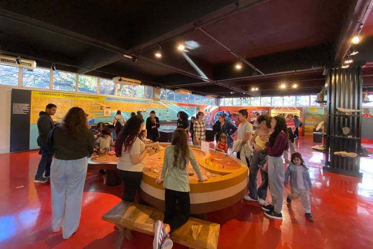 A imagem mostra um ambiente interno com várias pessoas interagindo em um espaço educativo. Há um grande balcão em formato circular no centro, onde algumas pessoas estão reunidas. O chão é vermelho e as paredes possuem painéis informativos. Ao fundo, há janelas que permitem a entrada de luz natural, e a atmosfera parece ser de aprendizado e diversão.