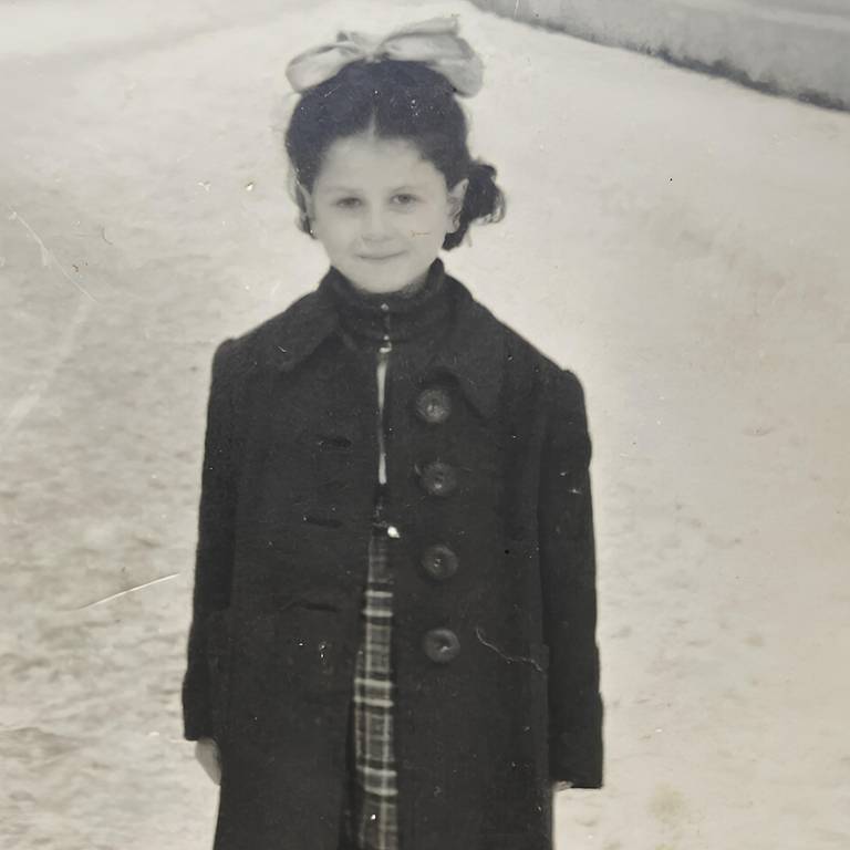 A imagem mostra uma menina em uma estrada, vestindo um casaco escuro e uma saia xadrez. Ela tem cabelo escuro preso em um coque e usa um laço grande na cabeça. O fundo é uma paisagem de estrada com vegetação ao lado, e a foto parece ser em preto e branco, sugerindo uma época passada.
