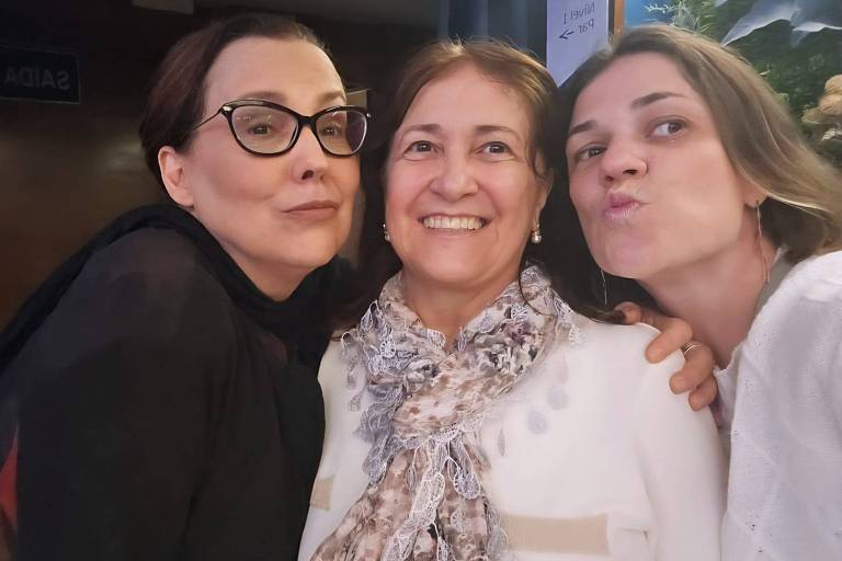 A imagem mostra três mulheres posando juntas em um ambiente interno. A mulher à esquerda usa óculos e tem cabelo escuro, fazendo uma expressão facial divertida. A mulher no centro sorri amplamente, vestindo uma blusa clara e um lenço ao redor do pescoço. A mulher à direita faz uma expressão de beijo, com cabelo claro e solto, vestindo uma blusa branca. O fundo é decorado com plantas.