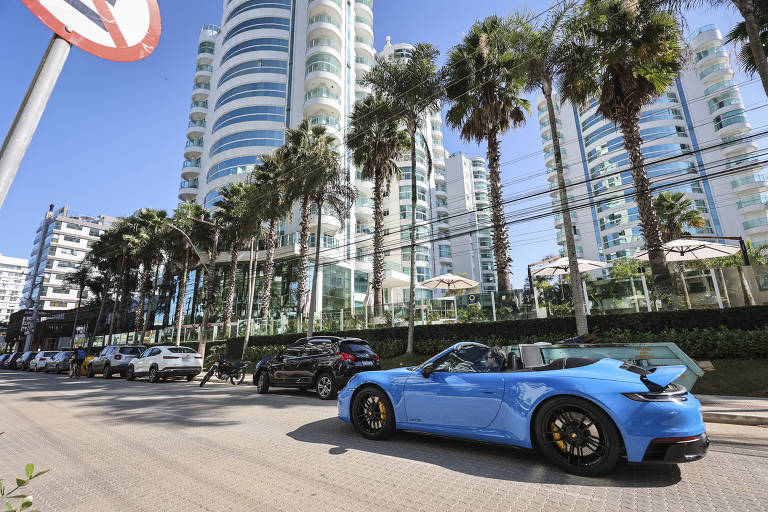 A imagem mostra uma rua urbana com vários carros estacionados. Em primeiro plano, há um carro esportivo azul. Ao fundo, há edifícios altos e modernos, com várias palmeiras ao longo da calçada. O céu está claro e ensolarado, e há uma placa de trânsito visível à esquerda.