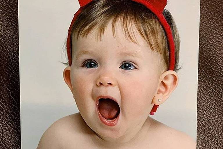 A imagem mostra um bebê sem camisa, com um grande laço vermelho na cabeça. O bebê está sorrindo e com a boca aberta, demonstrando alegria. Os olhos são claros e a pele é clara. A mão de um adulto, com unha pintada de rosa, está segurando a foto.
