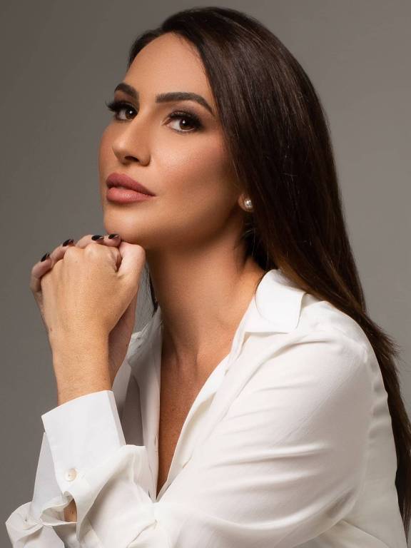 A imagem mostra uma mulher com cabelo longo e liso, usando uma camisa branca. Ela está posando com as mãos apoiadas no queixo, olhando para o lado. O fundo é neutro e desfocado, destacando a figura da mulher. A maquiagem é suave, com ênfase nos olhos e nos lábios.