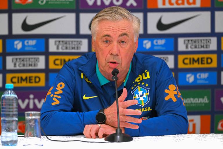 Um homem de cabelo grisalho, vestido com uma jaqueta azul da seleção brasileira, está sentado em uma mesa de coletiva de imprensa. Ele está falando e gesticulando com as mãos. Ao fundo, há um painel com logotipos de patrocinadores e a sigla da CBF. Na mesa, há uma garrafa de água e um copo com água.