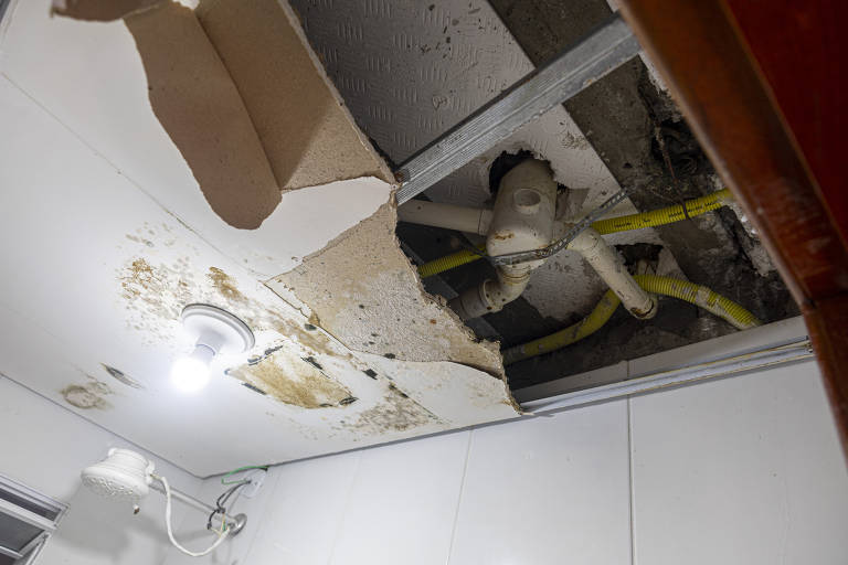 A imagem mostra um teto danificado, com uma parte do forro caída, revelando canos expostos e sinais de mofo. Há uma lâmpada acesa no teto e fios visíveis. O ambiente parece ser um banheiro ou uma área de serviço