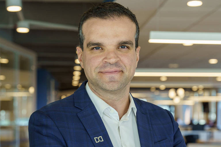 Um homem está posando para a foto em um ambiente de escritório moderno. Ele usa um terno azul escuro com uma camisa branca. O homem está com os braços cruzados e sorri levemente. Ao fundo, há mesas de trabalho e uma iluminação clara, sugerindo um espaço de trabalho colaborativo.
