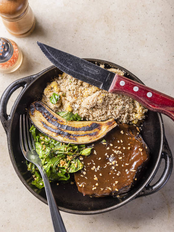 A imagem mostra um prato de comida servido em uma panela de ferro. O prato contém um pedaço de carne coberto com molho escuro, acompanhado de farofa, uma banana-da-terra grelhada e uma salada de folhas verdes. Um garfo e uma faca estão posicionados ao lado do prato. Ao fundo, há um saleiro e um tempero em pó.