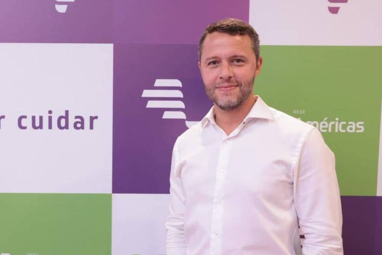 Um homem de camisa branca e calça escura está posicionado à direita da imagem, sorrindo. Ao fundo, há um painel com quadrados em cores roxa e verde, onde se lê a frase "Paixão por cuidar" e o logotipo da empresa "Fleury Américas".