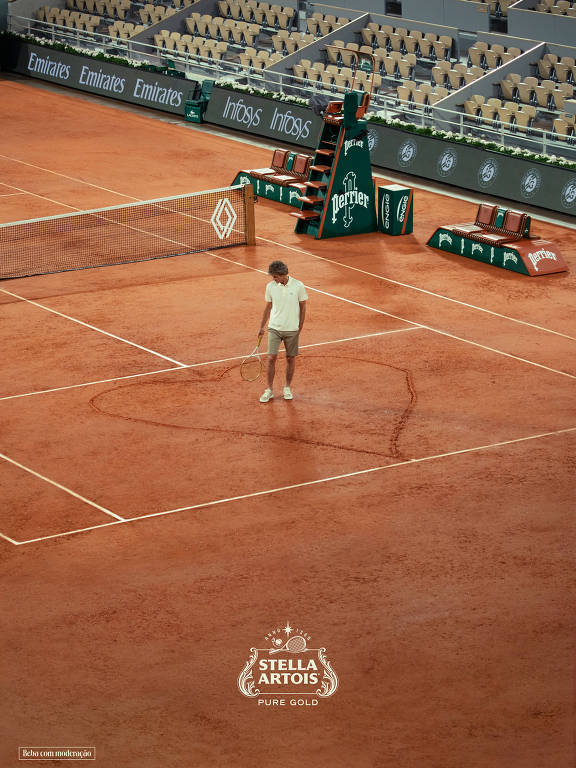 A imagem mostra um jogador de tênis em uma quadra de saibro, com um fundo de arquibancadas vazias. O jogador está vestido com uma camiseta branca e shorts, e parece estar se preparando para um saque. No fundo, há banners de patrocinadores e uma rede de tênis. O piso da quadra é de terra batida, característico de torneios de saibro.