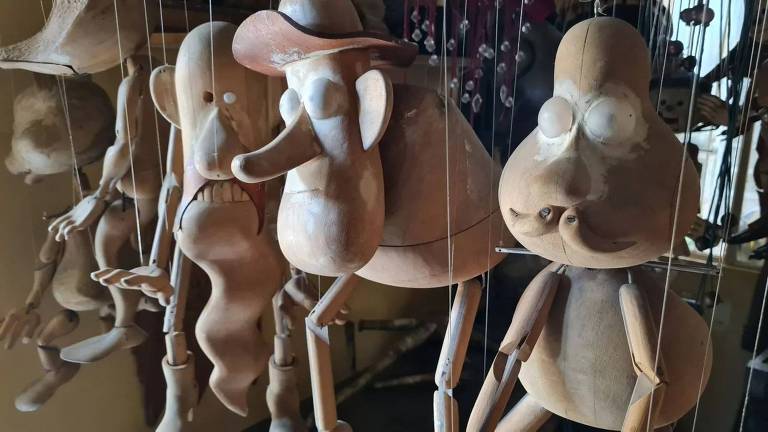 A imagem mostra várias marionetes de madeira penduradas. As marionetes têm formas distintas, incluindo um personagem com um chapéu, um rosto alongado e expressões variadas. Elas estão suspensas por fios, em um ambiente iluminado, com um fundo desfocado que sugere um espaço de trabalho ou uma loja de artesanato.