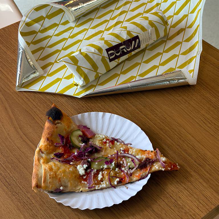 Uma fatia de pizza está em um prato branco, com coberturas visíveis como cebola roxa, picles e queijo. Ao fundo, há uma embalagem de papel com um padrão amarelo e branco, que parece ser de um restaurante chamado 'DORUM'. A mesa é de madeira clara.