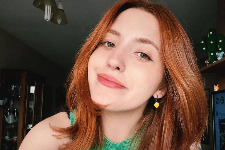 A imagem mostra uma mulher com cabelo ruivo e liso, usando uma blusa verde. Ela está sorrindo e olhando para a câmera, com um fundo desfocado que sugere um ambiente interno. A mulher tem brincos amarelos e algumas tatuagens visíveis nos braços. A iluminação é suave, destacando seu rosto e cabelo.