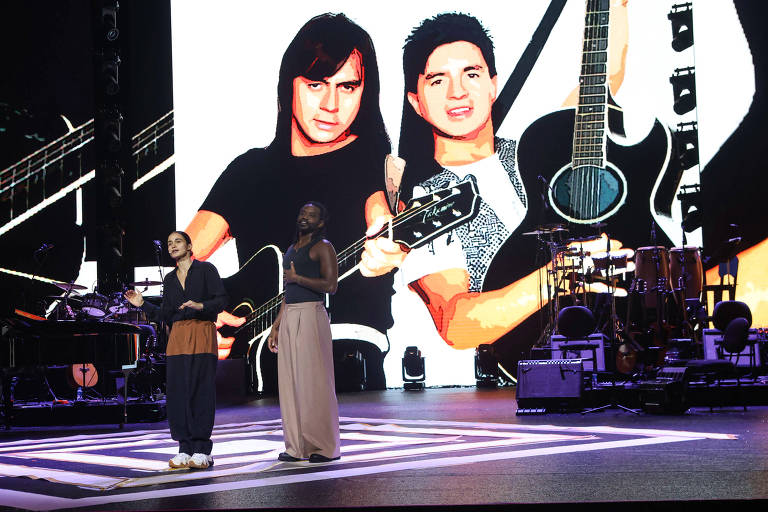 Duas pessoas estão se apresentando em um palco. Uma delas está vestida com uma blusa preta e calças largas, enquanto a outra usa uma blusa clara e calças longas. Ao fundo, há uma grande tela com a imagem de duas pessoas segurando guitarras. O ambiente é iluminado e há instrumentos musicais visíveis no palco.