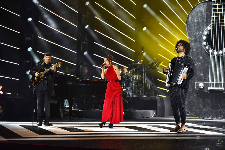 A imagem mostra três artistas se apresentando em um palco. À esquerda, um homem toca violão e usa um chapéu. No centro, uma mulher canta, vestindo um vestido vermelho. À direita, um homem toca sanfona e está descalço. O fundo do palco é iluminado com luzes amarelas e há uma grande representação de uma guitarra na parte de trás.