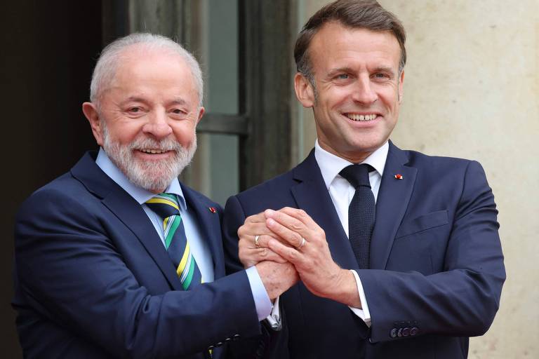 Dois homens estão de pé, sorrindo e apertando as mãos. O homem à esquerda tem cabelo grisalho e uma barba curta, vestindo um terno escuro com uma gravata verde e amarela. O homem à direita tem cabelo castanho claro e está usando um terno escuro com uma gravata preta. Ao fundo, há uma parede clara e uma janela.