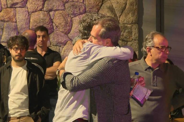 La imagen muestra a un grupo de personas en un evento, con dos hombres abrazándose. El fondo es un muro de piedra, y hay varias personas alrededor, algunas mirando a los dos hombres abrazándose. Los hombres visten de manera informal y el ambiente parece ser de camaradería.