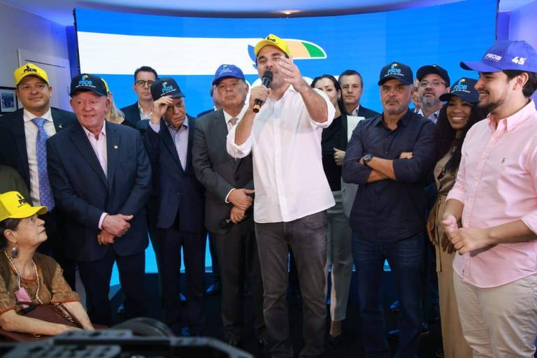 A imagem mostra um grupo de pessoas em um evento, todas usando bonés amarelos e azuis. No centro, um homem de camiseta branca está falando e gesticulando. Ao redor dele, há várias pessoas, incluindo homens e mulheres, algumas com expressões sorridentes. O fundo é uma tela com cores azul e verde, possivelmente representando uma marca ou evento. Uma mulher idosa está sentada à esquerda, enquanto outros participantes estão em pé, formando um círculo em torno do orador.