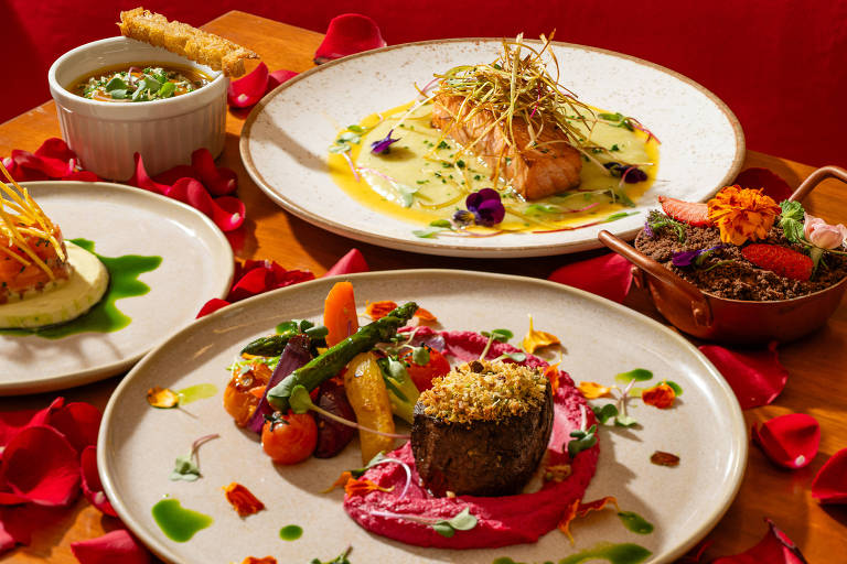 A imagem apresenta uma mesa decorada com pétalas de rosa. Em primeiro plano, há um prato com um filé de carne coberto com uma crosta dourada, acompanhado de purê de beterraba e vegetais coloridos. Ao fundo, um prato com salmão grelhado em um molho amarelo e decorado com ervas. Também há um recipiente com sopa e um prato com uma mistura de vegetais e flores comestíveis. A apresentação é sofisticada e colorida.