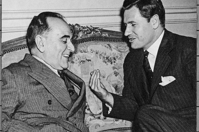 A imagem mostra dois homens conversando em um sofá. O homem à esquerda está sorrindo e usa um terno listrado, enquanto o homem à direita, também de terno, gesticula com a mão enquanto fala. O fundo é decorado com um padrão clássico.