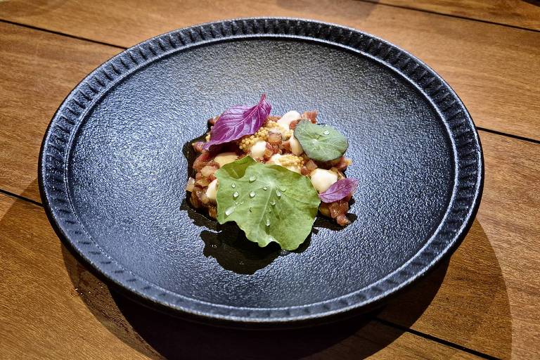 Um prato preto com uma apresentação de comida gourmet. No centro, há uma mistura de ingredientes que inclui pedaços de carne, ervas e flores comestíveis, como folhas verdes e pétalas roxas. O fundo é uma mesa de madeira, que contrasta com o prato escuro.