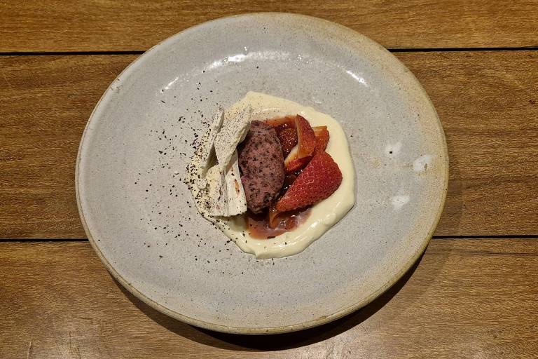 Um prato redondo e raso apresenta uma sobremesa composta por um creme claro, pedaços de morango, uma porção de massa ou merengue e uma cobertura de pó escuro, possivelmente chocolate ou café. O fundo do prato é de um tom neutro, e a mesa é de madeira, com um acabamento liso.
