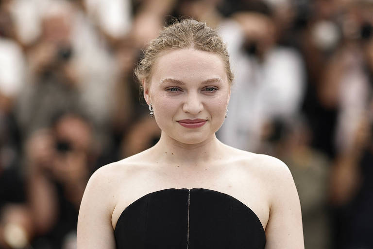 Uma mulher está em um evento de tapete vermelho, usando um vestido preto sem mangas. Ela tem cabelo preso e está sorrindo levemente. Ao fundo, há várias pessoas, algumas segurando câmeras, sugerindo que é uma ocasião de grande atenção da mídia.
