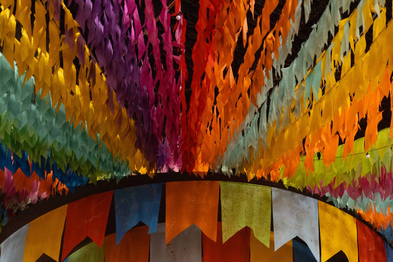 A imagem mostra uma decoração vibrante com tiras de papel colorido penduradas em um ambiente. As tiras estão dispostas em um padrão radial, criando um efeito visual dinâmico. As cores incluem roxo, laranja, verde, azul e amarelo, formando um arco-íris de tonalidades. A parte inferior da imagem apresenta bandeirinhas de papel em várias cores, complementando a decoração.
