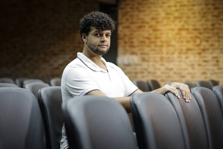 Um homem com cabelo cacheado e barba está sentado em um auditório. Ele usa uma camiseta branca com detalhes escuros e está posicionado ao lado de cadeiras de plástico. O fundo é composto por uma parede de tijolos expostos e a iluminação é suave, criando um ambiente acolhedor.