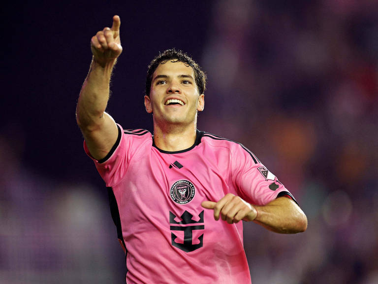 Um jogador de futebol está em campo, vestindo uma camisa rosa com detalhes em preto. Leo Afonso está sorrindo e apontando para cima com a mão direita, enquanto a multidão ao fundo parece animada. O ambiente é de um estádio, com luzes e torcedores visíveis.