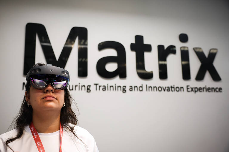 Uma pessoa está usando óculos de realidade aumentada e está em frente a uma parede com a palavra 'Matrix' em destaque. Abaixo do nome, há a frase 'Manufacturing Training and Innovation Experience'. A pessoa parece concentrada e está vestindo uma camiseta branca com um crachá vermelho.