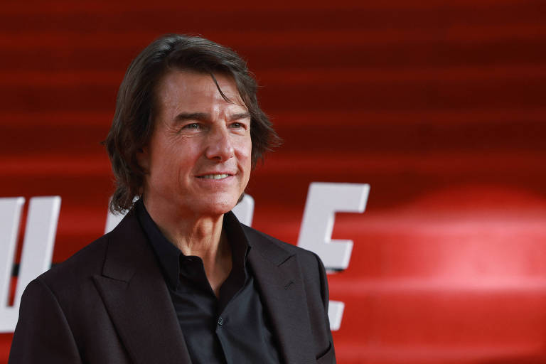 Um homem com cabelo castanho e liso, usando um terno escuro, está posando em um evento de cinema. Ao fundo, há uma parede vermelha com letras brancas que parecem ser parte de uma promoção ou cartaz. O homem está sorrindo levemente e parece estar em um ambiente de tapete vermelho.