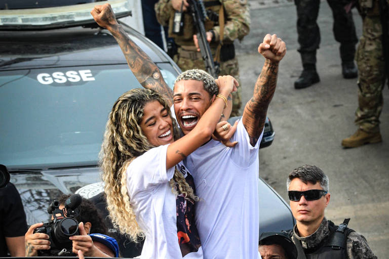 A imagem mostra um casal sorridente em cima de um carro, ambos vestindo camisetas brancas. Eles estão abraçados e levantando os braços em sinal de celebração. Ao redor, uma multidão de pessoas está levantando os celulares para registrar o momento. Ao fundo, há policiais e veículos de segurança, indicando um ambiente de festa ou evento público.