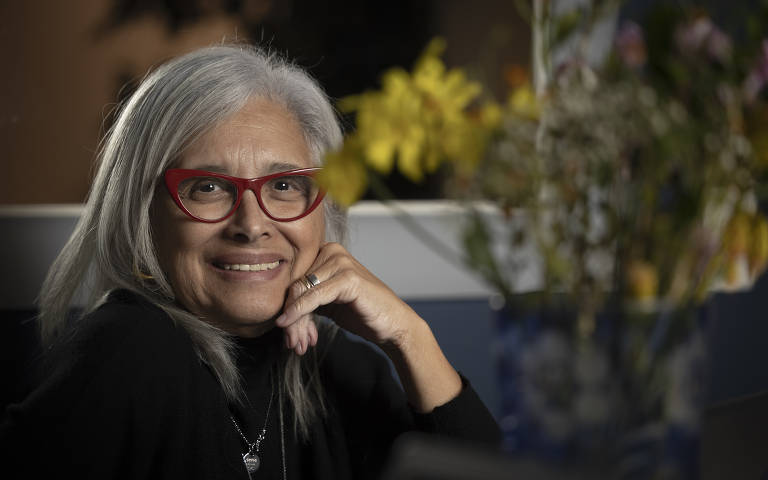 A imagem mostra uma mulher sorrindo, com cabelos grisalhos e longos, usando óculos vermelhos. Ela está apoiando o queixo com a mão e vestindo uma blusa preta. Ao fundo, há um vaso com flores coloridas, que adiciona um toque de cor à cena. A iluminação é suave, destacando seu sorriso e a expressão amigável.