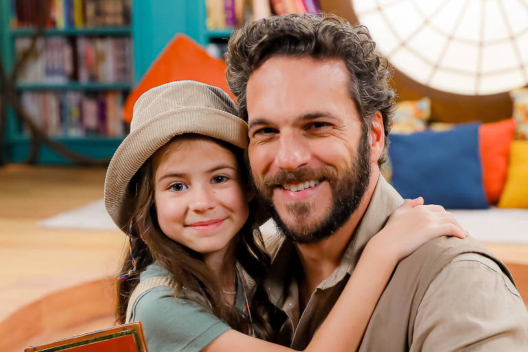 A imagem mostra um homem e uma menina sentados em uma mesa de madeira em um ambiente interno. O homem, com barba e cabelo cacheado, está sorrindo e segurando a menina, que usa um chapéu e sorri de volta. A menina segura um livro intitulado 'Viagem ao Centro da Terra', que apresenta uma ilustração de um globo terrestre na capa. Ao fundo, há estantes de livros e almofadas coloridas dispostas em um espaço aconchegante.
