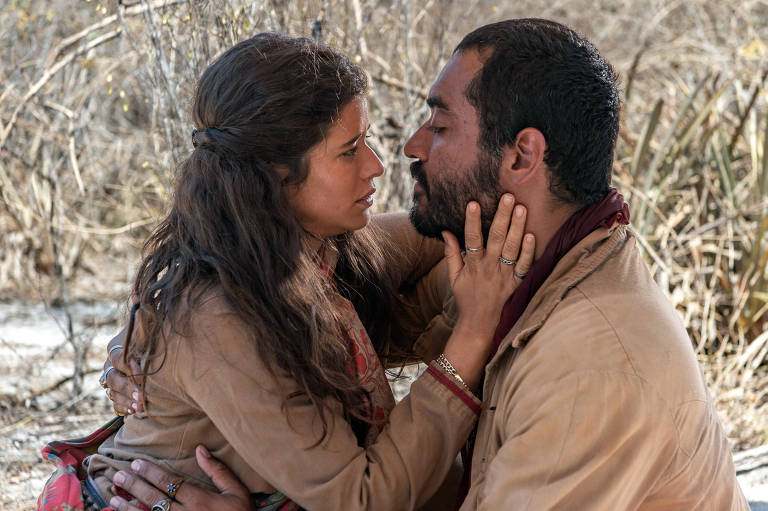 A imagem mostra um casal em um momento íntimo, com os rostos próximos e expressões emocionais intensas. A mulher tem cabelo castanho e usa uma blusa clara, enquanto o homem tem barba e veste uma camisa escura. O fundo é composto por vegetação seca, sugerindo um ambiente natural e árido.