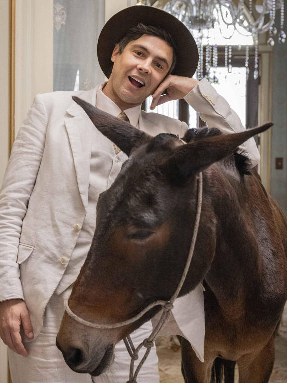 Um homem vestido com um terno claro e chapéu está posando ao lado de um burro em um ambiente interno. O homem sorri e parece estar se divertindo, enquanto o burro está com a cabeça baixa, próximo ao homem. O fundo apresenta uma decoração elegante, com um lustre visível e paredes decoradas.