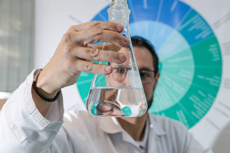 Um cientista está segurando um frasco de laboratório com líquido transparente. Ele está usando um jaleco branco e óculos. Ao fundo, há um gráfico colorido que parece representar dados científicos ou informações sobre biologia. A iluminação é clara e o ambiente parece ser um laboratório.