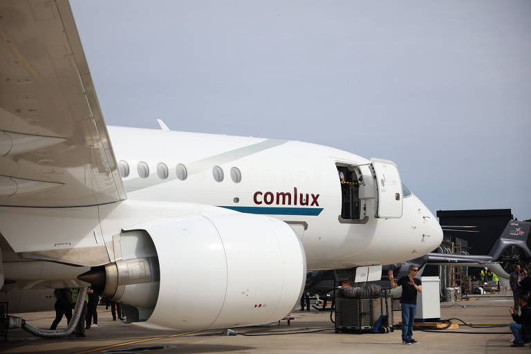 A imagem mostra a parte frontal de um avião da Comlux, com um motor visível e a porta de embarque aberta. Algumas pessoas estão ao redor do avião, e o céu está parcialmente nublado.