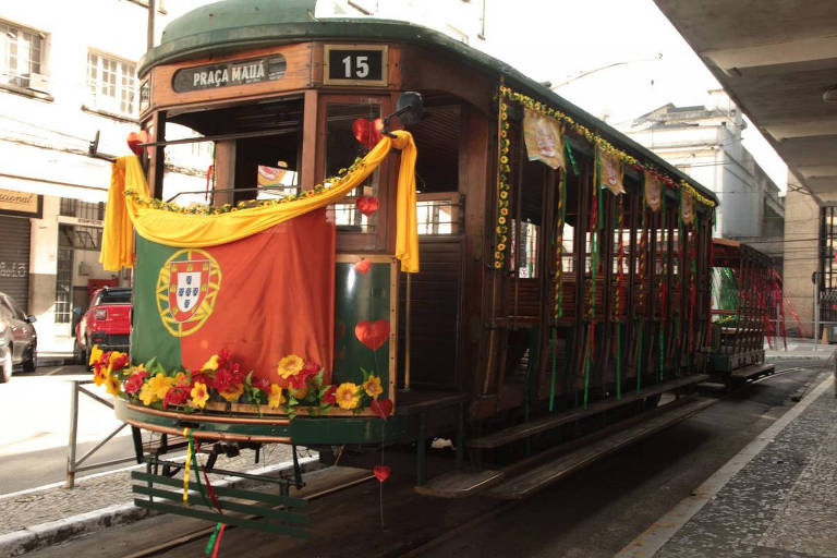 A imagem mostra um bondinho decorado, com flores e fitas coloridas. Na parte frontal, há uma bandeira de Portugal e o número 15. O bondinho está estacionado em uma rua, com prédios ao fundo e um calçamento de pedras.