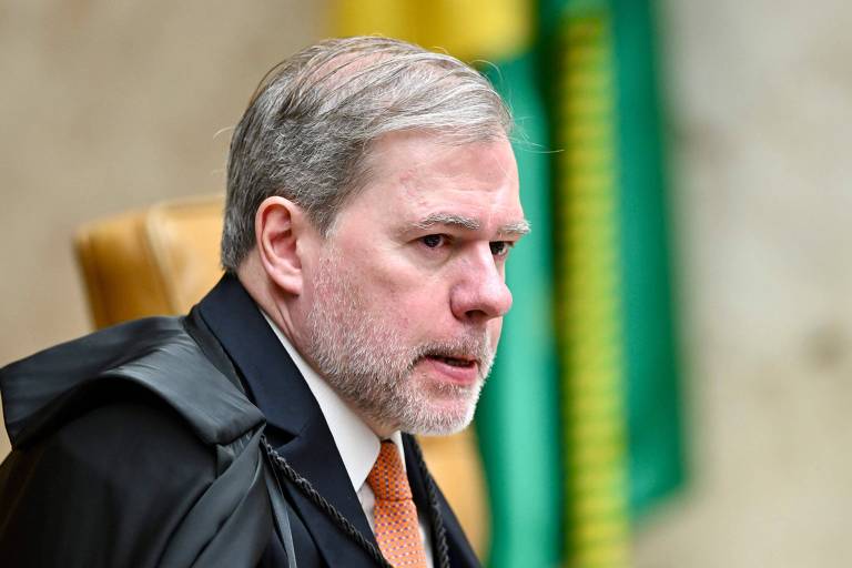 Een man met grijs haar en een baard, gekleed in een zwarte toga en een wit overhemd met een oranje stropdas. Hij zit op een stoel, kijkt opzij en heeft een serieuze blik. Op de achtergrond zijn een Braziliaanse vlag en een groene doek te zien.