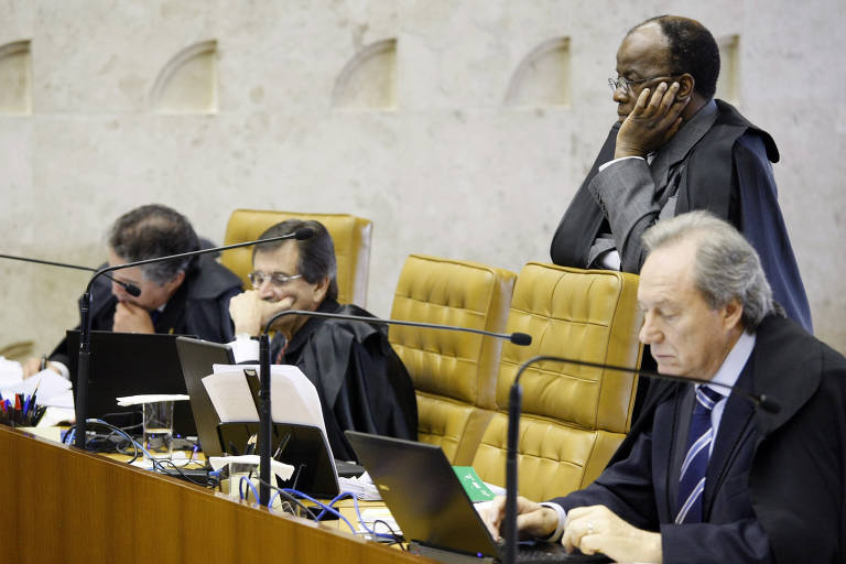 A imagem mostra uma sessão do Supremo Tribunal Federal do Brasil, com quatro ministros sentados à mesa. Eles estão usando togas e estão concentrados em seus laptops. Um dos ministros está em pé, com uma expressão pensativa. O ambiente é formal, com uma mesa de madeira e um fundo claro.