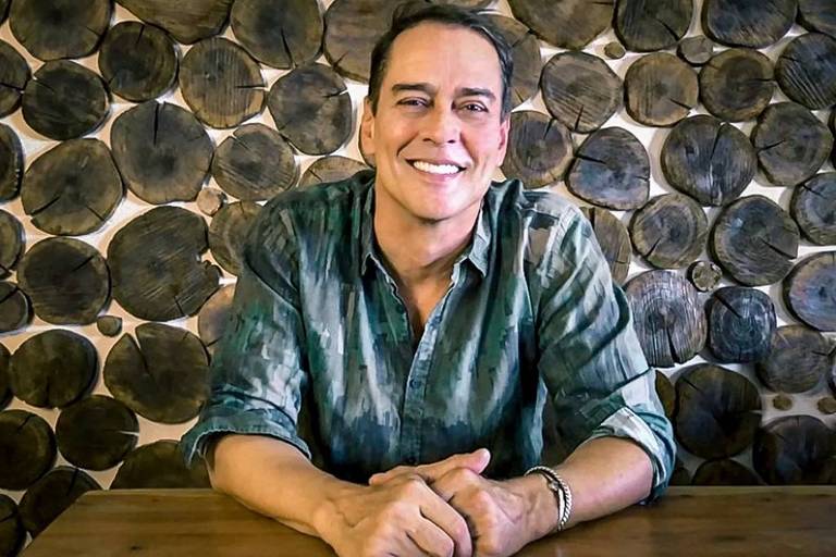 Um homem sorridente está sentado à mesa, com as mãos apoiadas. Ele usa uma camisa de estampa camuflada em tons de verde e azul. Ao fundo, há uma parede decorada com troncos de madeira dispostos em círculos. A iluminação é suave, destacando o sorriso do homem.