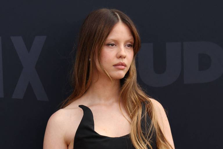 Uma jovem mulher está posando em um evento, vestindo um vestido preto justo. O vestido tem um detalhe assimétrico em um dos ombros. Ela tem cabelos longos e soltos, e está em pé com uma expressão séria, com as mãos unidas à frente do corpo. O fundo é escuro, com a palavra 'NETFLIX' visível em letras grandes.
