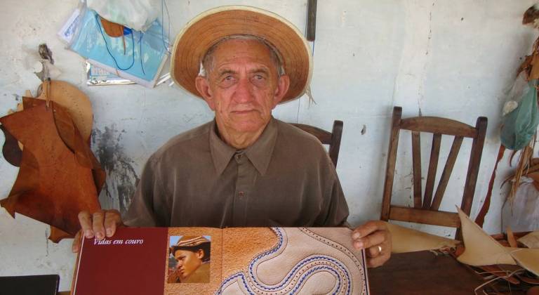 Um homem idoso, com um chapéu de palha, está sentado em uma cadeira de madeira. Ele segura um livro aberto que mostra imagens relacionadas ao trabalho com couro. Ao fundo, há pedaços de couro pendurados na parede e uma mesa de madeira. O ambiente parece rústico e simples, com uma parede clara e alguns objetos de trabalho visíveis.