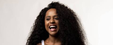 Duda Santos *** Local Caption *** A imagem mostra uma mulher com cabelo cacheado e volumoso, usando uma camiseta branca sem mangas e calças largas de jeans. Ela está sorrindo amplamente, com as mãos na cintura, em um fundo claro e neutro.