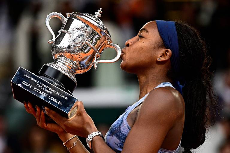 Uma jogadora de tênis está beijando um troféu. Ela usa uma blusa azul e tem cabelo longo e solto. O troféu é prateado e possui uma base preta com inscrições. O fundo da imagem mostra uma multidão, sugerindo que a cena ocorre em um evento esportivo.