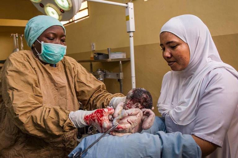 A imagem mostra duas profissionais de saúde em um ambiente hospitalar. Uma delas, vestindo um avental e máscara, está segurando um recém-nascido, que ainda está coberto de sangue. A outra profissional, usando um hijab e também com máscara, observa e parece estar ajudando. O ambiente é iluminado e há equipamentos médicos visíveis ao fundo.