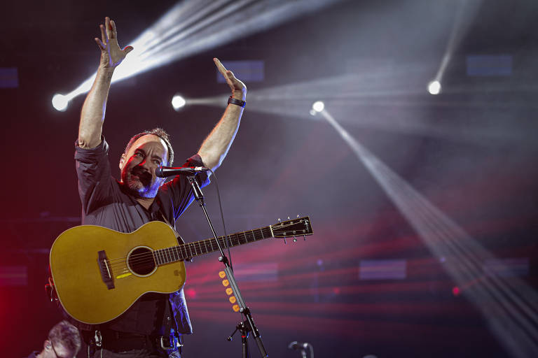 Um artista está se apresentando ao vivo em um palco, segurando um violão. Ele está com os braços levantados, expressando emoção. O fundo é iluminado por luzes brilhantes, criando um ambiente vibrante e dinâmico.